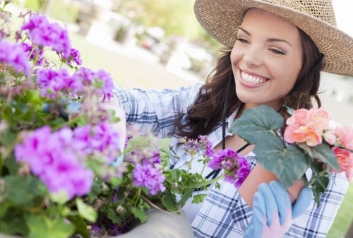 A woman with long brown hair, wearing a wide-brimmed straw hat and light blue gardening gloves, is smiling while tending to a garden filled with vibrant purple and pink flowers in a residential outdoor space. The foreground features densely planted flowering bushes with rich green leaves, and the background shows a well-maintained lawn and a blurred view of garden elements in a sunny environment, suggesting a tidy, landscaped yard in Shepherds Bush. The scene captures the natural textures of the lush foliage, colorful blooms, and the smooth surface of a gardening table or pot, reflecting careful gardening care typical of local outdoor maintenance services. The bright daylight highlights the vivid colors and healthy plant growth, emphasizing a neat and inviting garden setting suitable for professional gardening and lawn care by Lawn Mowing Shepherds Bush.