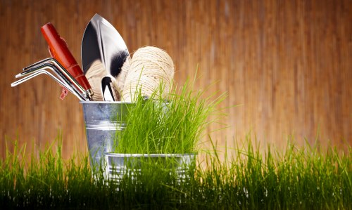 A close-up image of a collection of gardening tools placed inside a small metal bucket, sitting on a lush, green grass surface. The tools include a pair of hedge shears, a small hand rake, a trowel, and a red-handled garden fork, with a ball of twine positioned next to them. The background features a warm, wooden surface, suggesting a garden shed or wooden fencing, with natural lighting highlighting the vibrant green grass and metallic textures of the tools. The scene reflects a well-organized gardening setup suitable for lawn care and general outdoor maintenance in Shepherds Bush, aligning with services offered by Lawn Mowing Shepherds Bush.