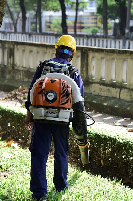 A gardener dressed in an orange high-visibility uniform, including a cap, is watering a lush, flowering shrub with a hose, standing on a well-maintained lawn. The garden features a variety of plants, 